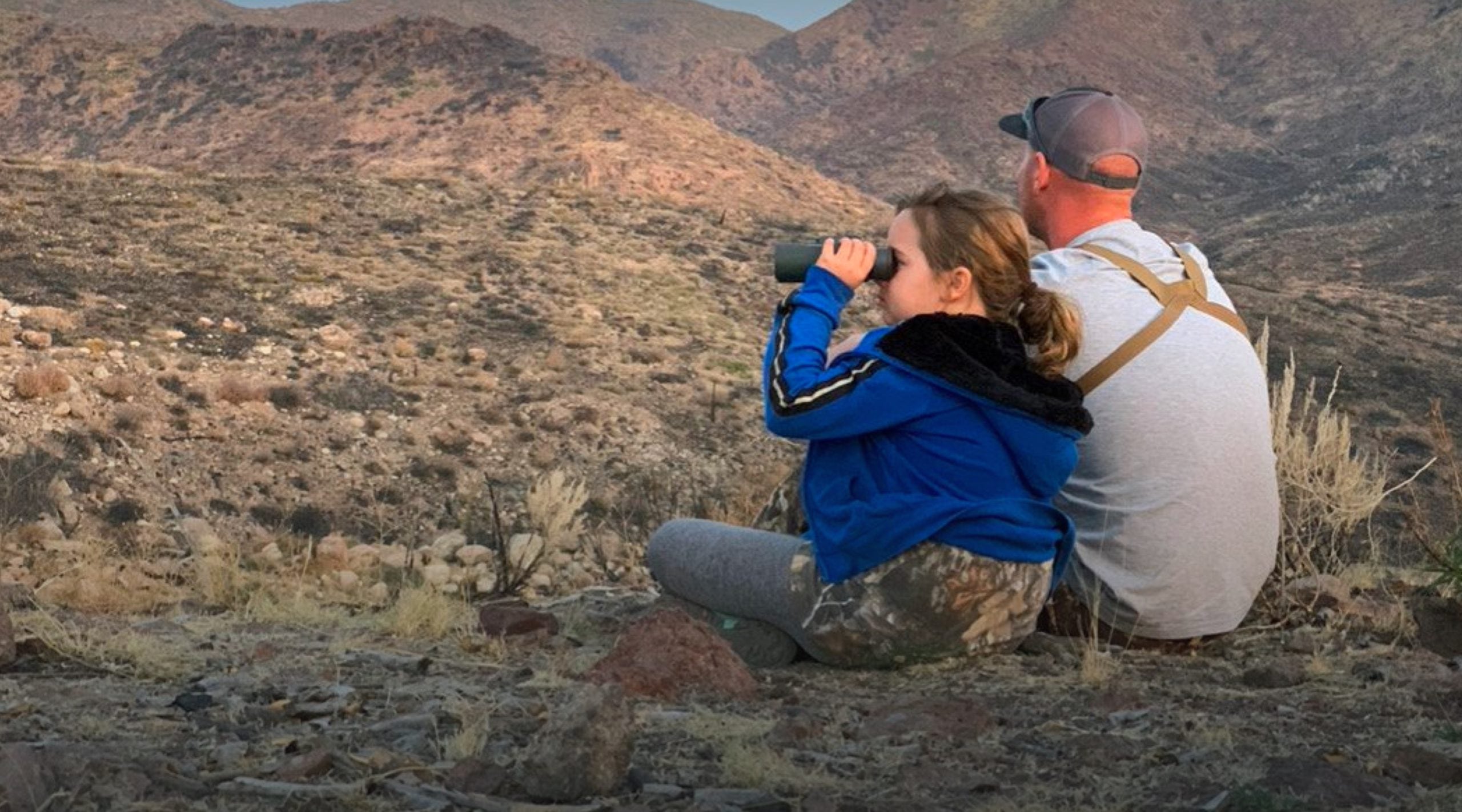 A little girl looks through binoculars as she sits with her dad looking over a Nevada landscape on a scouting trip.