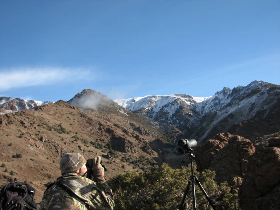 Nevada hunter looks through binoculars and has spotting scope set up next to him on snow-dusted mountain top