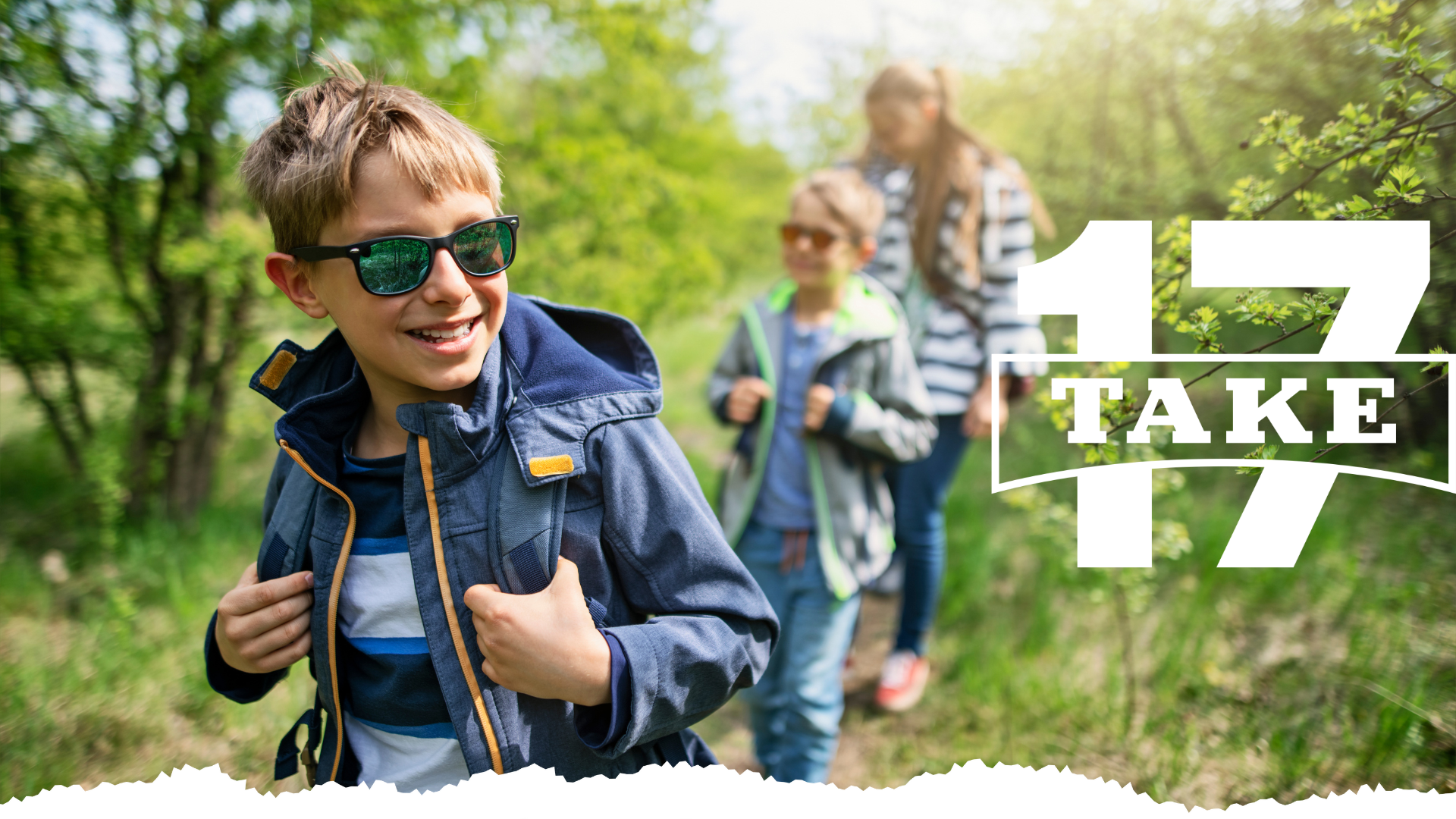 Young boy with sunglasses on smiling as he hikes with his family following behind him.